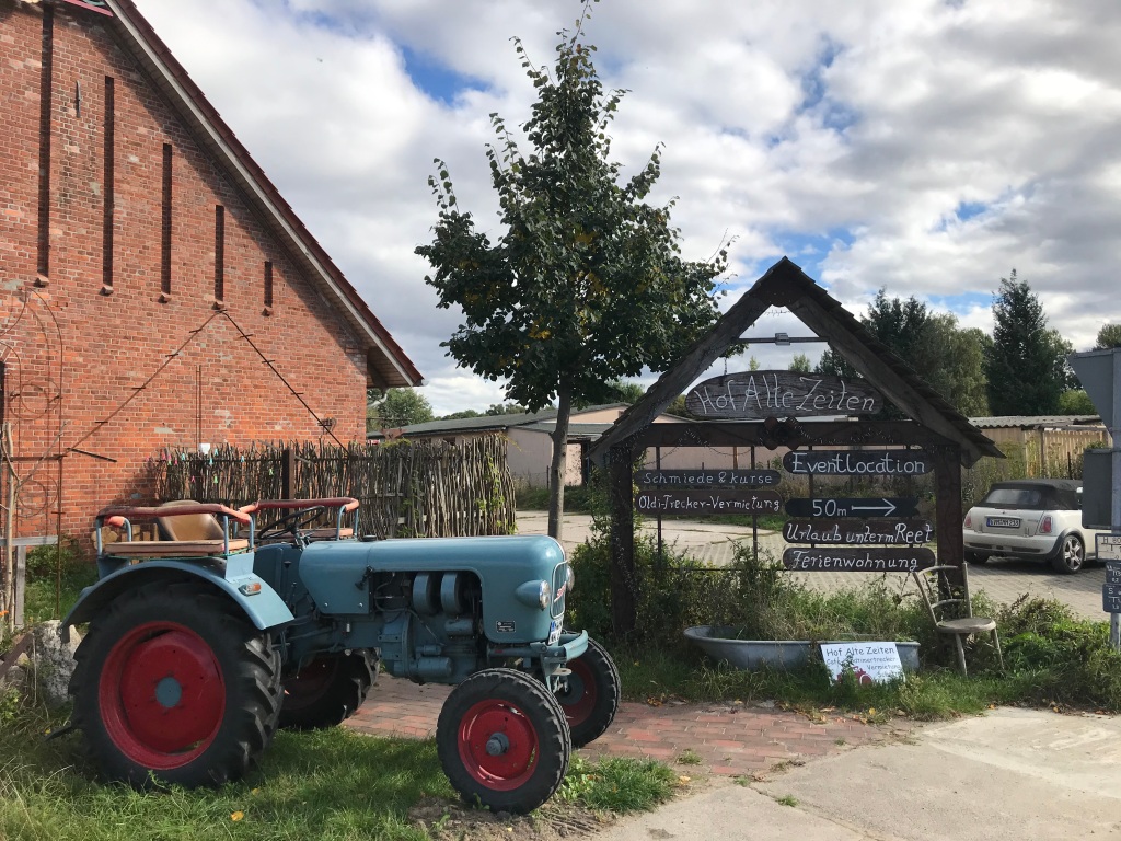 Dargestellt ist ein Traktor aus den 1950er Jahren vor einem landwirtschaftlichem Gebäude und der Werbung für Urlaub auf dem Bauernhof
